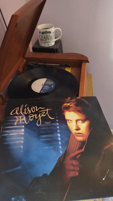 Record player with a 12" black vinyl record.
In the foreground is an album cover showing a softly lit Alison Moyet in front of window blinds with light filtering through.
Behind the player is a mug of coffee on a speaker.