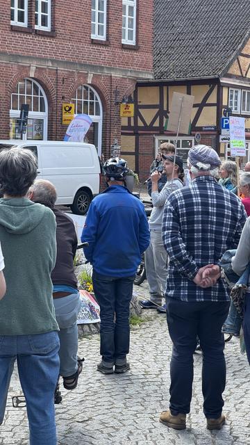 A group of people gathered in a public space, listening to a speaker holding a microphone. Some attendees are taking notes or holding signs. The scene features a mix of casual clothing and a backdrop of brick buildings and banners.