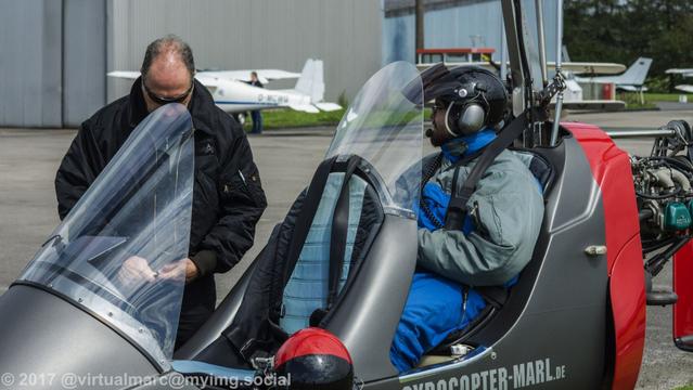 A photo of a Gyrocopter on the ground. The pilot is preparing his suit. I'm sitting in full gear in the passenger seat behind the pilot.