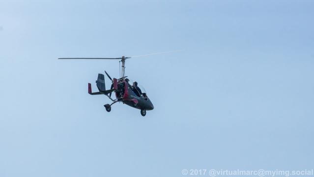 A photo of a gyrocoper in the air flying from the ground against blue sky. You can see the pilot in the front and me as a passenger in the back.