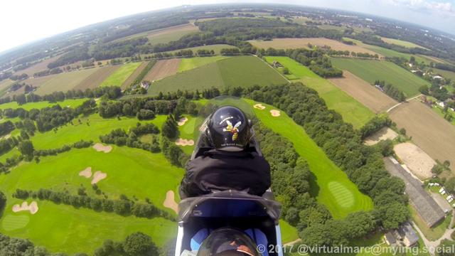 A photo of a GoPro Hero 3 camera mounted below the top rotor of a Gyrocopter. You can see part of the gyrocopter with me in the back and the pilot in the front, from behind. We are currently flying over a Golf course near the airport in Marl.