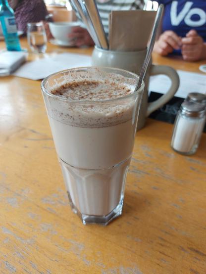 Im Vordergrund ein Glas mit Chai Latte dahinter Speisekarten Salzstreuer, weitere Getränke und verschwommen einige Menschen. Eine Person trägt ein T-Shirt mit der Aufschrift "Volt"
