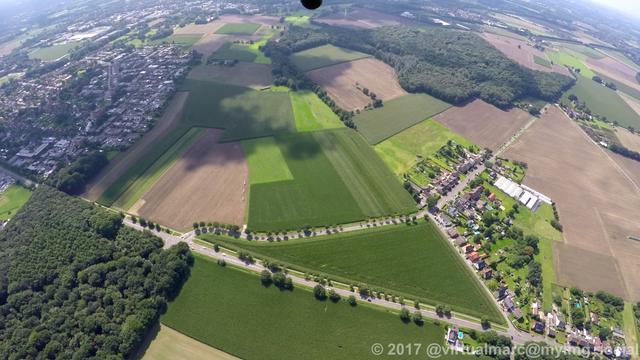 A photo of agricultural fields and some wood from above. This is in Herten. A street and some houses can be seen.