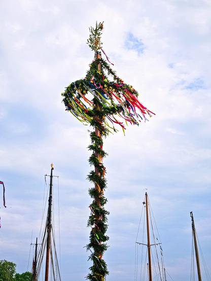 In the centre of the photo is a maypole. This is a long, narrow pole wrapped with fir branches. A kind of crown is attached to the upper end, also wrapped in fir branches. Colourful strips of fabric blow in the wind from the crown. More branches can be seen on top of the crown. This is a custom for 1 May. Ship masts can be seen to the left and right of the maypole. All against a cloudy sky.