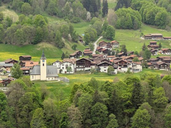 A small town, nestled in the mountains, lots of white and brown alpine style buildings, clustered around a yellowy/white church tower