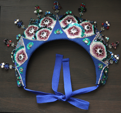 A blue crescent shaped headdress on a dark background. It has a ribbon at the end tied in an upside down bow. The headdress is decorated with sequins and mirrors and beads in a pattern that looks like peacock feathers.