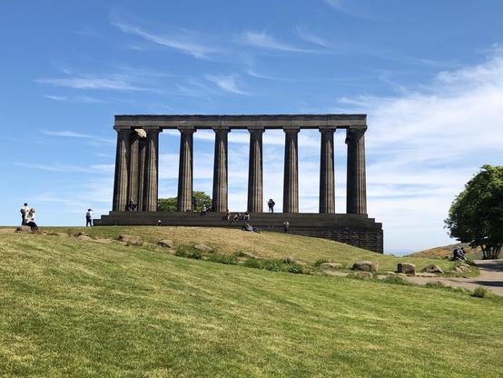 The image shows the National Monument of Scotland, located on Calton Hill in Edinburgh. It is an unfinished monument originally intended to be a replica of the Parthenon in Athens as a memorial to the Scottish soldiers and sailors who died fighting in the Napoleonic Wars. The monument features tall classical columns, and the scene is captured on a sunny day with clear blue skies. People are seen around the monument, some walking and others sitting on the grass, enjoying the outdoors.