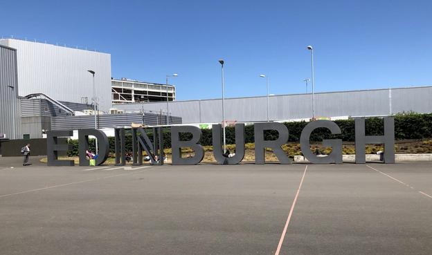 The image features large standalone letters spelling out "EDINBURGH," likely located in a public space near an industrial or commercial building, as suggested by the structure in the background. There are a few individuals near the letters, one of whom appears to be taking a photo of the installation. The setting seems to be a parking lot or open plaza area, and it is a sunny day with clear skies.