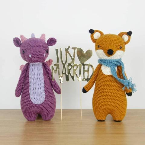 A picture of a crocheted purple dragon and a crocheted orange fox with a blue scarf. They're holding a cake topper that says 'just married'.