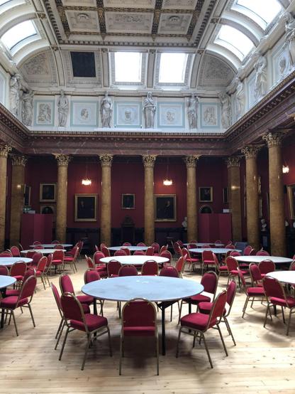This image depicts an elegant and ornate hall with a classical architectural style. The room is characterized by a series of large red columns, an intricately decorated ceiling with detailed moldings and frescoes, and a high ceiling with clerestory windows that allow natural light to illuminate the space. The walls are adorned with dark red tones and several framed portraits. Round tables, each surrounded by red chairs, are arranged throughout the room, suggesting that the space is used for events or gatherings. The floor is wooden, adding warmth to the grandeur of the hall. Overall, the image showcases a sophisticated and historically rich interior, likely used for formal events or assemblies.