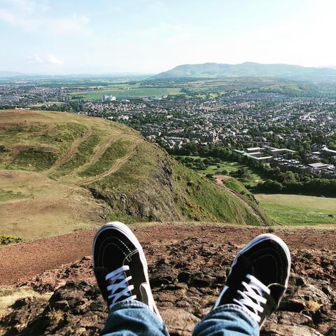 The image shows a beautiful panoramic view from a high vantage point over a lush, green landscape and a town. In the foreground, a person's legs are extended with their feet wearing sneakers, dangling over a rocky edge, suggesting they are sitting and enjoying the view from a high location. The visible paths across the greenery indicate popular hiking or walking trails. In the distance, there is a town spread out with various buildings, and beyond the town, a range of hills or mountains can be seen under a clear sky. This setting might perhaps be somewhere known for its scenic hikes and views.