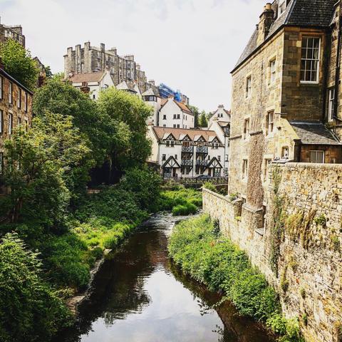 This image shows a beautiful, historic urban scene featuring old buildings along the sides of a narrow river. The architecture is quite varied, displaying elements of traditional Tudor-style timber framing on some buildings with their distinctive white and black exterior patterns. Other structures are constructed from stone, adding to the rustic, old-world charm of the setting. The scenery is lush with green vegetation lining the riverbank and filling the space between the buildings, enhancing the quaint and picturesque atmosphere. The overall look suggests this could be a location in an older European city, possibly in the United Kingdom.