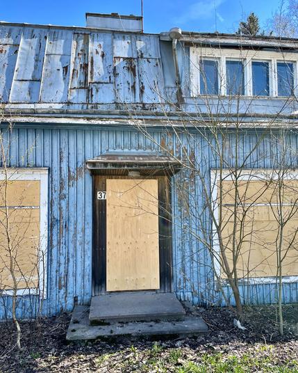 Abandoned blue house with peeling paint, boarded windows, and a wooden door near the entrance, surrounded by overgrown bushes.