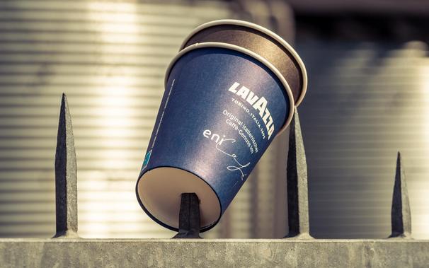 Ein blauer Kaffeebecher der Marke Lavazza steckt aufgespießt auf einem Zaunpfahl mit spitzen Metallzacken. Der Becher wirkt verloren oder absichtlich platziert, was ein ungewöhnliches Bild erzeugt. Im Hintergrund sind unscharfe industrielle Elemente zu erkennen, die der Szene eine raue Atmosphäre verleihen.