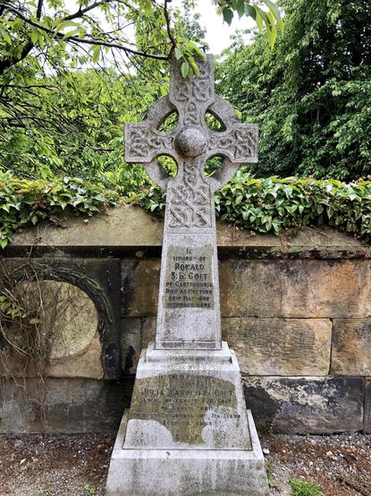 This image shows an ornate stone Celtic cross gravestone. The cross features intricate knotwork designs, a common motif in Celtic art, emphasizing circular forms and interlacing patterns. The inscription on the gravestone commemorates individuals named Ronald S.F. Colt and Jean Cardin F.H. Colt. It mentions that Ronald S.F. Colt died on 22 May 1936 and was interred at the location of the gravestone. Jean died on 10 May 1938 at the age of 71 years. The stone cross stands before a wall covered with some ivy and is surrounded by verdant vegetation, likely located in a serene cemetery setting.
