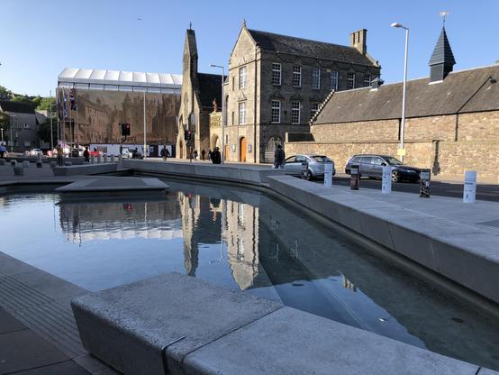 This image features a modern urban scene with a variety of architectural styles. It shows a public square with a contemporary water feature in the foreground, composed of shallow rectangular pools that reflect the surrounding buildings. The architecture in the background includes a mix of old and new buildings. Notably, there's an old stone building with traditional design elements next to a modern structure with a large, tent-like roof possibly used for events or as a cover for a public space.

There are a few people walking and engaging in regular city activities, and cars parked along the side of the street. The street and the public area are paved, and the photo is taken on a sunny day, contributing to the pleasant, vibrant atmosphere of the scene. The flags and traffic lights add to the urban environment detail, suggesting this is likely a significant or central part of the city.