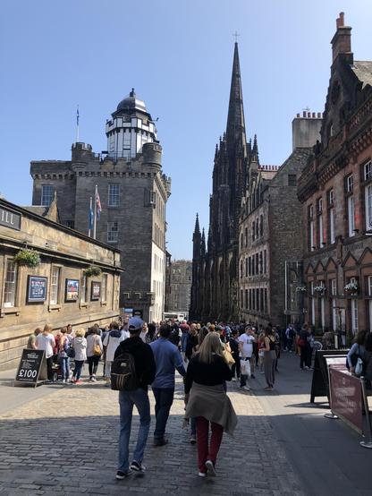 This image captures a bustling street scene in what appears to be Edinburgh, Scotland. The architecture and the visible landmarks, such as the distinctive tower on the left resembling the Camera Obscura and World of Illusions, and the historic Gothic spire of the Hub on the right which is a key part of the city's skyline, suggest it's taken on or near the Royal Mile. This location is famous for its historical and cultural significance in the heart of Edinburgh's Old Town.

The street is paved with cobblestones and lined with traditional, multi-story stone buildings displaying a variety of flags. In the foreground, numerous tourists and locals are seen walking and perhaps shopping or exploring the area. The weather is sunny, casting sharp shadows and contributing to the vivid atmosphere of what looks like a busy day in the city.