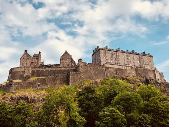 The image shows the iconic Edinburgh Castle, situated atop Castle Rock in the city of Edinburgh, Scotland. The castle is a prominent historical structure known for its significant role in Scottish history. The architecture visible in the image includes sturdy stone walls and several ancient buildings with turrets and a larger, more rectangular structure. The castle overlooks the lush green area surrounding it, and the sky above is bright with scattered clouds. This setting highlights the castle's strategic position and picturesque appeal, making it a popular tourist attraction.