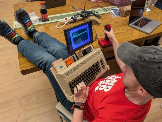 Madtixx sitzt im Saarbrücker Hackspace am Tisch, die Beine auf den Tisch gelegt, mit einem C64 auf dem Schoß und einem Eingabegerät in der Hand. Am C64 sind diverse Dinger eingesteckt, von denen der Autor dieses Beschreibungstextes keine Ahnung hat, vor allem ist aber ein Bildschirm eingesteckt, sodass sich das Gerät wie ein Laptop nutzen lässt.