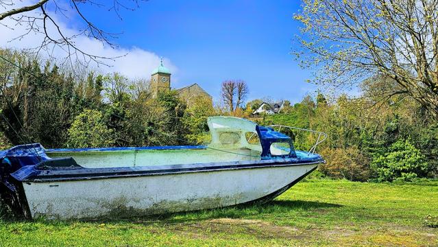 A weathered white and blue boat rests on green grass in a field, with trees and a church steeple visible in the background under a bright blue sky.