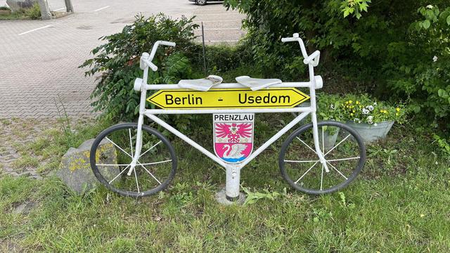 Ein skurriles Fahrrad als Fahrradschild mit Richtungspfeilen, die auf "Berlin" und "Usedom" zeigen, mit einem Wappen mit der Aufschrift "Prenzlau". Das Fahrrad ist weiß gestrichen und auf grasbewachsenem Boden in der Nähe eines Blumentopfs ausgestellt.