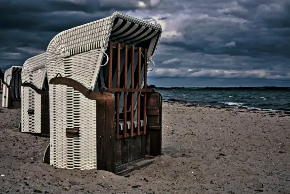 Ein noch (oder wieder?) verschlossener Strandkorb steht auf einem Sandstrand, dahinter sind zwei weitere Körbe zu sehen. Rechts läuft die Ostsee an den Strand, die See ist kabbelig. 
Der Himmel ist von dunklen Wolken verhangen. Regen kündigt sich an.