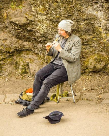 The image features a man seated on a folding chair against a rugged stone backdrop, likely outdoors. He is playing a tin whistle and wears a casual, layered outfit consisting of a gray tweed blazer, a gray beanie, black pants, and dark shoes. Beside him on the ground are a few personal items, including a backpack and a green towel, with a cap placed upside down, likely for collecting tips from passersby, suggesting he might be a street performer. The natural setting and his relaxed demeanor give the scene a serene and artistic feel.