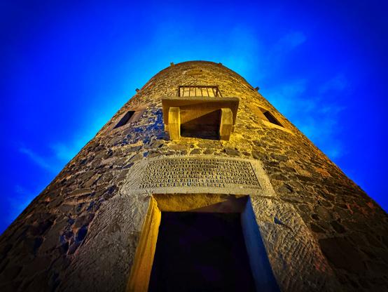 A tall stone tower is viewed from below against a deep blue sky. The tower features a small balcony and a stone inscription near its entrance, illuminated by warm lighting.