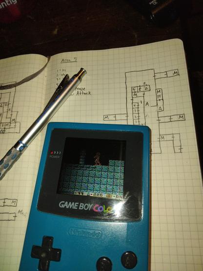 Photo of a Game Boy Color with Metroid II playing, sitting on a graph paper notepad with a hand drawn map.
