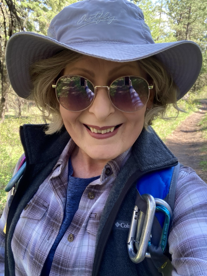 On a trail that’s visible in the background, along with pine forest, an older woman smiles for the camera. She’s wearing a purple and white flannel shirt that’s open in the front, a blue shirt beneath that. There’s also a charcoal, fleece vest. On the blue straps of her pack are several carabiners. She’s wearing tinted sunglasses, a beige, wide-brimmed hat over her blonde hair.
