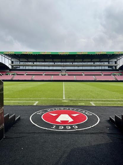 This image shows the interior of a football stadium. The view is from behind one of the goals, looking across the pitch towards the opposite stand. Prominently featured in the foreground is a logo on the ground marking the entrance to the pitch, which reads "FC Midtjylland 1999," indicating this is the home stadium of the Danish football club FC Midtjylland, which was founded in 1999. The stadium features green seating with the name "Faxe Kondi" displayed across the opposite stand, suggesting a sponsorship or partnership. The field itself is lush and green, marked for football play.