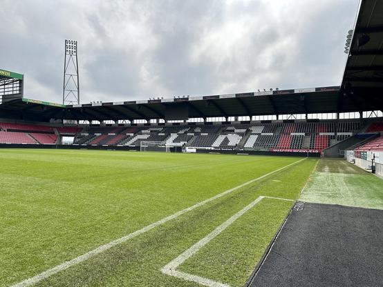 This image shows an empty soccer stadium with a well-maintained grass pitch. The stadium features multiple tiers of seating, which are mostly unoccupied, and the spectator sections are accented with design elements in black, white, and red. There is a tall floodlight tower visible on the left, and part of the stadium structure includes cantilevered roofs over the seating areas. This setting shows an overcast sky and some hints of sunshine, creating a serene and somewhat dramatic atmosphere, typifying a professional sports venue awaiting an event or match.