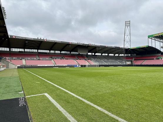 This image shows a football stadium. It features a well-maintained grass pitch with white boundary lines and some writing near the edge. The stadium has several sections with red seating, and there are covered stands to shelter spectators. The facility is equipped with large floodlights for illumination during night matches or poor visibility conditions. There are also various advertisement banners visible around the stadium. The stadium appears empty, suggesting that the photo was taken during a non-match time or possibly a private visit.