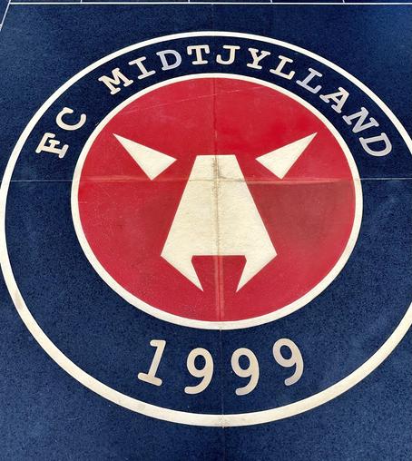 This image features the logo of FC Midtjylland, a professional football club in Denmark. The logo includes a red and white emblem with a stylized representation of a wolf, surrounded by a dark blue circle that contains the text "FC MIDTJYLLAND" and the year "1999", indicating the club was established in that year. The club's colors and logo are prominently displayed.