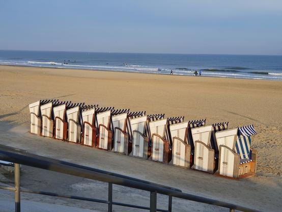 Das Foto zeigt dicht aneinandergereihte Strandkörbe. Sie stehen am Rande des Sandstrandes, zu dem eine Treppe hinunterführt. Der Strand und die Körbe sind von der Sonne beschienen; die etwas weiter entfernte blaugrau schimmernde Nordsee ist ruhig und hat lediglich kleine auslaufende weiße Wellen. Es sind nur wenige Menschen direkt am Wasser zu sehen.
