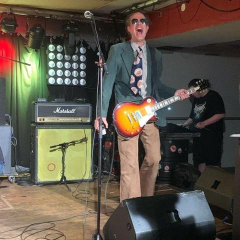 Bild zeigt einen Menschen mit E-Gitarre (Les Paul) auf einer Bühne. Er trägt eine Sonnenbrille, ein grünes Sacko, eine 70er-Jahre-Krawatte und ein gemustertes Hemd. Er hat den Mund wie bei einem Schrei geöffnet. Im Hintergrund neben ihm ist ein Marshall-Verstärker samt Box zu sehen.