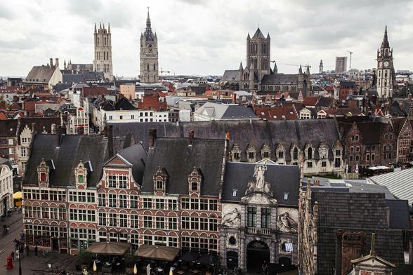 A view from a high building in the centre of a European city with a medieval history. It's an overcast day and there's a feeling that there's been rain in the recent past given the damp look to the buildings we can see. We're looking over the tops of roofs of four and five storey buildings, many with very tall, steep roofs, some with stepped gables typical of northern European countries. Four church towers are spaced across the city's skyline, two with clock faces on them.