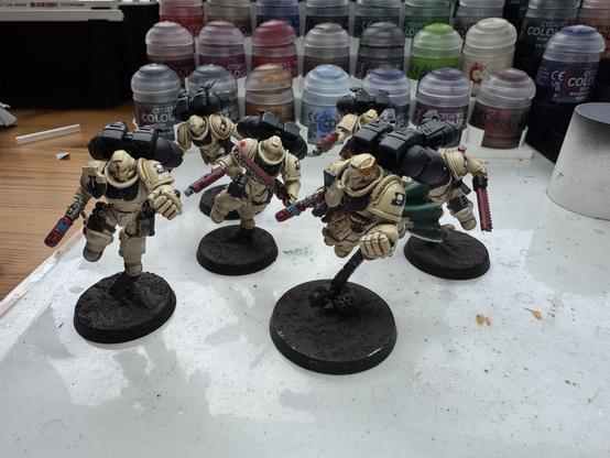 6 Warhammer Miniaturen vor einem Regal voller Farbtöpfchen