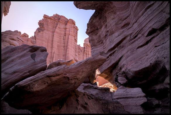the view from a canyon, in the foreground are big orange rocks in the shadow, through a rock window bright orange rocks in the background are visible