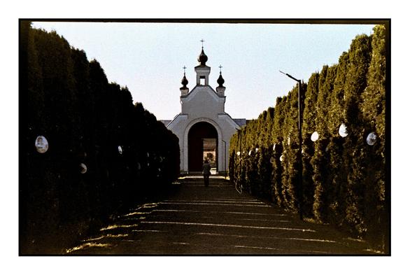 A person walking down a tree-lined path toward a small white chapel, captured on expired Fujicolor 200 film, with visible color shifts and grain adding a vintage, nostalgic feel.