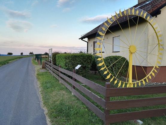 Neben der Wetterwarte östlich der gesuchten Ortschaft steht ein gelber, zahnrad- oder flammenähnlicher Kranz, daneben führt ein asphaltierter Weg nach hinten. Einige Objekte rechts des Weges sind nur undeutlich zu erkennen.
(Foto: Joddel1123, CC-BY-SA 4.0, Wikimedia Commons, 2016)