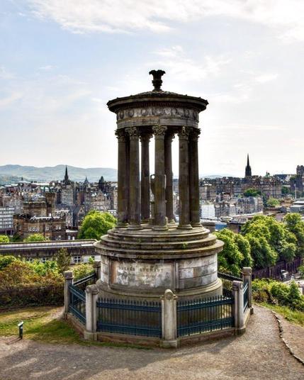 This image shows the Dugald Stewart Monument, which is a memorial to the Scottish philosopher Dugald Stewart (1753–1828). It is situated on Calton Hill overlooking the city of Edinburgh, Scotland. The monument features a circular temple design, styled in a Neoclassical manner, based on the Choragic Monument of Lysicrates in Athens. This setting offers a panoramic view over the city, encompassing both its historic and more modern parts, nestled amid the natural landscapes surrounding it. The monument and its location are popular for providing one of the iconic vistas of Edinburgh, blending cultural heritage and natural beauty.