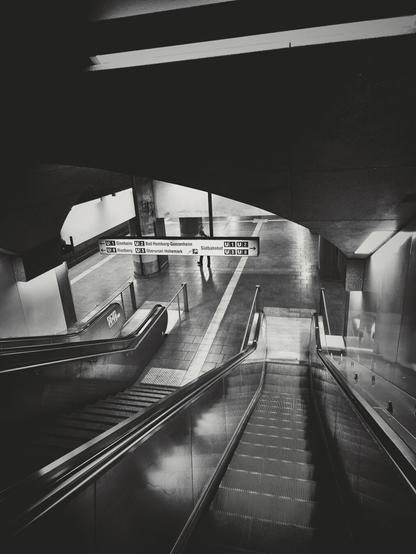 The image shows a black and white view of an escalator system. There are two escalators descending from the viewer, separated by a central divider. In the background, another escalator or stairs appear to be ascending. Overhead signage is visible, including the sign with arrows pointing left and right and text in German, reads "Ginnheim", "Riedberg", "Bad Homburg", "Südbahnhof", etc. The area around the escalators is tiled, and there are reflections on the surface of the descending escalators. A person is visible in the background. Lighting illuminates parts of the area, casting shadows.