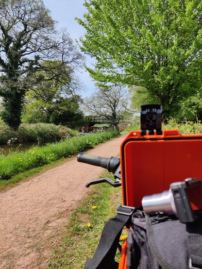 On the tow-path of a canal. In the background, a decorated canal barge is pulled up under a short, steel fabricated bridge with stone abutments. In the foreground (and slightly unfocussed) is a pushbike handlebar with an orange plastic case open in front of it. Visible inside the case is a small camera with a extended zoom lens, and a nylon carrying bag - presumably full of other equipment (I think it was my sound gear).