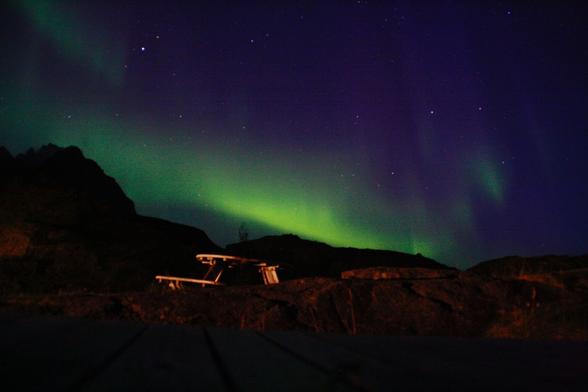 Nighttime landscape showing green aurora borealis streaks across a dark sky with stars. Silhouetted mountains form the horizon. A wooden picnic table is visible in the foreground, dimly lit.