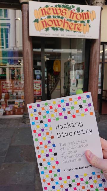 A paperback book, its cover a colourful grid of inter-connected multi-coloured little squares and titled "Hacking Diversity.  The politics of inclusion in open technology cultures" by Christina Dunbar-Hester, is held up in front of the entrance to a shop.

The sign above the doorway reads "News From Nowhere.  Liverpool's radical and community bookshop"