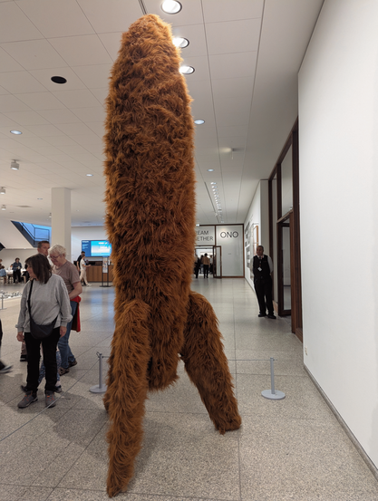 Sculpture First Spaceship on Venus on exhibit in a museum foyer. A tall rocket on three legs is coated in fluffy brown fur all over