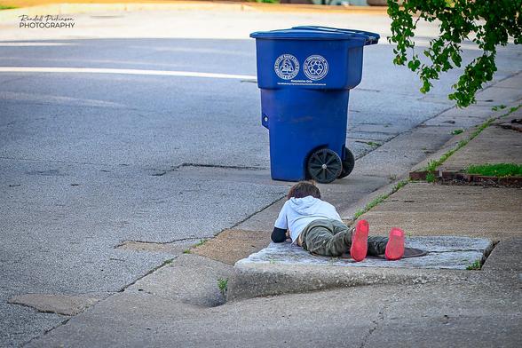 A young boy, maybe 8 or so years old, lays on top of a storm sewer inlet structure at the curb of a street, looking or playing with something out of view in front of him.  A blue plastic trash can sits in the street 10 feet beyond him.