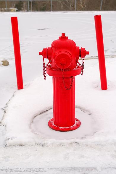 A photograph of a bright red fire hydrant standing out against the snowy landscape, flanked by two red bollards.