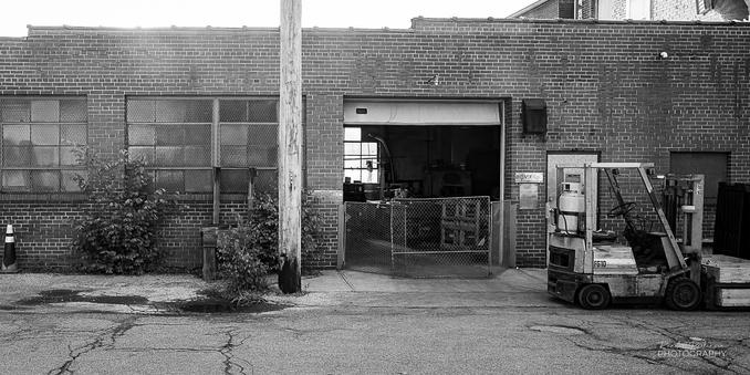 A street view of a metal plating company.  The one story brick building has large, multi-pane windows and an open garage door with a free standing fence/gate which one could assume is to keep dogs and cats from wondering inside.  A forklift sits on the street at the curb outside the garage entrance.